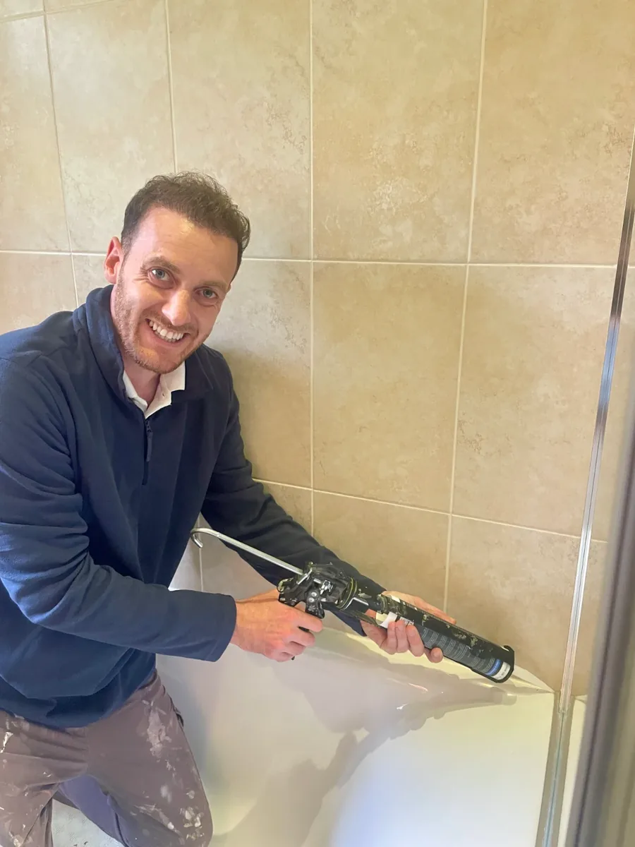 Mr Mastic applicator kneeling beside a white bath, sealing the joint where the bath meets beige wall tiles